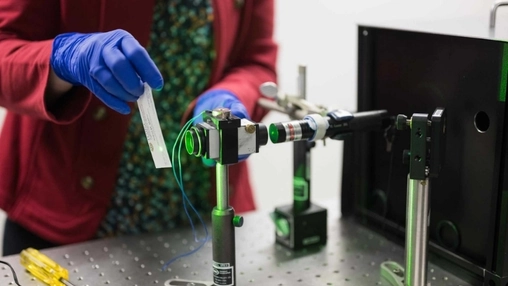 Researcher in blue gloves handling a clear sample near a green laser optics rig on a perforated lab bench