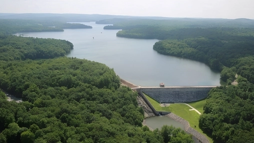 Drone photograph of a forested reservoir and concrete spillway dam with surrounding woodland and hills