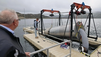 A cylindrical submarine device suspended by an orange gantry crane over a floating dock on a calm reservoir