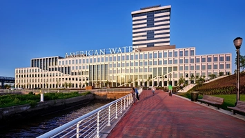 American Water headquarters with illuminated roof signage beside a brick waterfront walkway under a blue sky