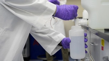 A laboratory researcher in a white coat and purple gloves filling a plastic bottle from a dispensing unit