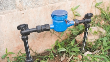 Blue water meter with black pipes and valves mounted near a stone wall with green plants at the base
